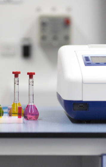 A laboratory benchtop with a row of five volumetric flasks containing blue, purple, green, yellow, and pink liquids, next to a white and blue electronic laboratory instrument A laboratory benchtop with a row of five volumetric flasks containing blue, purple, green, yellow, and pink liquids, next to a white and blue electronic laboratory instrument